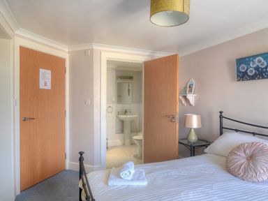 A bedroom with a bed, round pillow, bedside table with lamp, two doors, and an en suite bathroom at 9 Maritime House in West Bay
