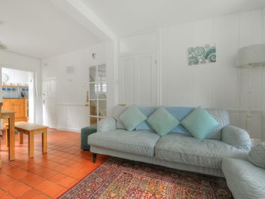 A living room with a light blue sofa with cushions a standing lamp a multicolored rug wooden dining table with benches and a view into a kitchen at Monmouth Cottage in Lyme Regis