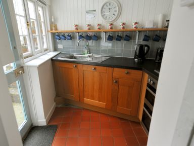 A kitchen corner with wooden cabinets a black countertop a stainless steel sink and mugs hanging under a shelf at Monmouth Cottage in Lyme Regis