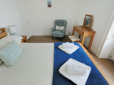 A bedroom with a bed towels a blue patterned chair a wooden dressing table and a framed picture at Monmouth Cottage in Lyme Regis