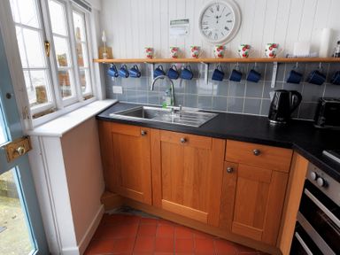 A kitchen with wooden cabinets a stainless steel sink blue mugs hanging under a wooden shelf and a wall clock at Monmouth Cottage in Lyme Regis