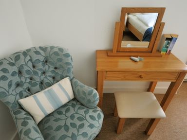 A floral armchair with a cushion next to a wooden dressing table with a mirror and a stool at Monmouth Cottage in Lyme Regis