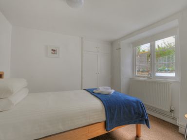 A bedroom with a bed covered with white sheets and blue blanket next to a window and built in wardrobe at Monmouth Cottage in Lyme Regis