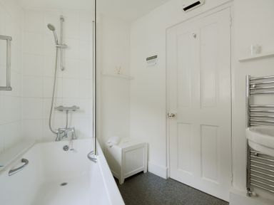 A bathroom with a bathtub and shower, a white door, a small storage box, and a towel radiator at Monmouth Cottage in Lyme Regis