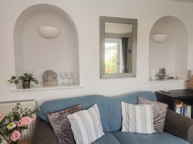 A living room with a blue covered sofa with cushions a mirror on the wall and decorative items on shelves at Bay View in Lyme Regis