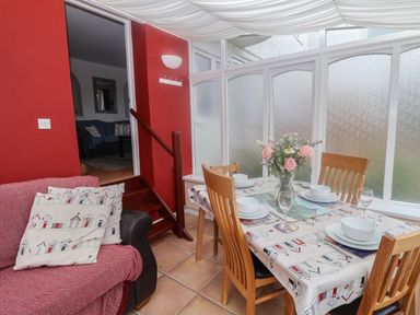A dining area with a table set for four with a flower vase and wooden chairs next to a red wall with an armchair and stairs at Bay View in Lyme Regis