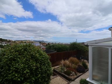 A garden with a large bush and various plants next to a patio and a conservatory with houses and hills in the background at Bay View in Lyme Regis
