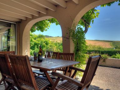 A patio with wooden table and chairs with a bottle and glasses under arches with a view of fields at Willows in Charmouth