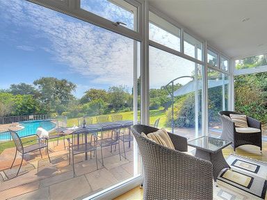 A sunroom with two wicker chairs a glass table and sliding glass doors leading to a patio with a dining table and chairs by a pool at Somerhill in Lyme Regis