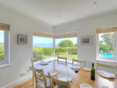 A dining room with a wooden table set with plates wine glasses and a bottle of wine near large windows overlooking a garden and pool at Somerhill in Lyme Regis