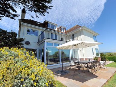 An outdoor patio with a table and chairs under a large umbrella next to a two-story house with balconies and large windows at Somerhill in Lyme Regis