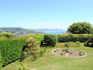 A garden with a circular flower bed and hedge with a coastal view at Somerhill in Lyme Regis