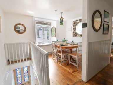 A dining area with a round wooden table, four chairs set with plates and glasses, a window with blinds, mirrors on the walls, and a staircase with white railing at Coombe Street Cottage Lyme Regis