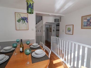 A dining area with a wooden table set with plates bowls and green glasses a bottle on the table white chairs white railing and framed artwork on the walls at Coombe Street Cottage Lyme Regis