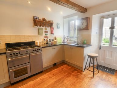 A kitchen with a stove oven, countertop with kettle and containers, sink under a window, stool and door at Coombe Street Cottage Lyme Regis