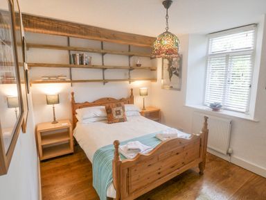 A bedroom with a wooden bedframe two bedside tables with lamps wall shelves and a window with blinds at Coombe Street Cottage in Lyme Regis