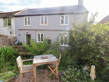 A garden with wooden chairs and table in front of a grey house with white framed windows at Coombe Street Cottage in Lyme Regis