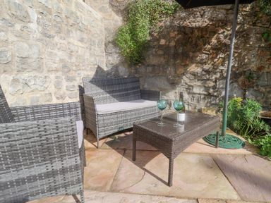 An outdoor patio area with gray wicker furniture a table with wine glasses and a bottle near stone walls at Coombe Street Cottage in Lyme Regis