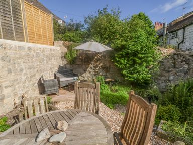 A small outdoor garden area with wooden table and chairs stone walls and wicker seating at Coombe Street Cottage in Lyme Regis