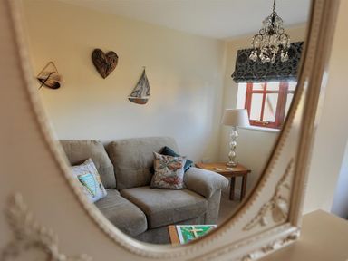A reflection in a mirror showing a beige couch with patterned cushions near a wooden side table with a lamp and a window with a patterned curtain at 28 Barnes Meadow in Uplyme