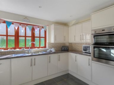 A kitchen with white cabinets a double oven a microwave a toaster and a window with colorful bunting at 28 Barnes Meadow in Uplyme