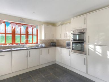 A kitchen with white cabinets a double oven a microwave a toaster and a window with colorful bunting at 28 Barnes Meadow in Uplyme