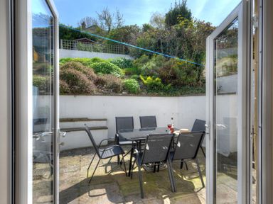 A patio with a glass table and six black chairs with a garden on a slope behind at Ivy Cottage in Beer