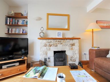 A living room with a stone fireplace a television on a wooden stand shelves with books a floor lamp and a coffee table with magazines and a cup at Ivy Cottage in Beer