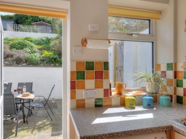 A kitchen countertop with colorful tiles and containers next to a window and a door opening to an outdoor seating area at Ivy Cottage in Beer