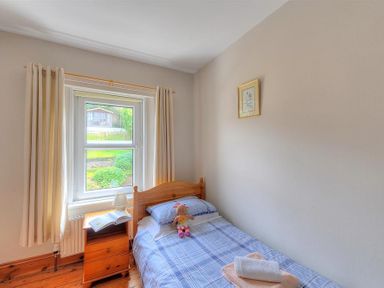A single bed with plaid bedding a wooden bedside table with a lamp and an open book in a bedroom at Ivy Cottage in Beer