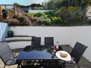 An outdoor patio with a black glass table and six black chairs surrounded by white walls and greenery at Ivy Cottage in Beer