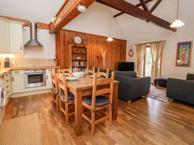 A kitchen and living room with a wooden dining table and chairs a bookshelf sofas a television and a kitchen area at 3 Home Farm South in Rousdon