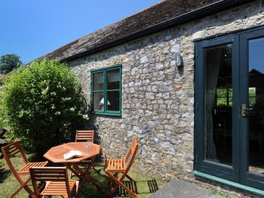 An outdoor patio area with a wooden table and chairs next to a stone wall with a window and glass doors at 3 Home Farm South in Rousdon