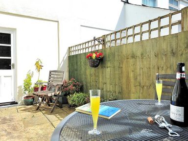 A patio area with wooden chairs potted plants a bottle of prosecco two glasses and a book on a metal table at Vine Cottage in Charmouth