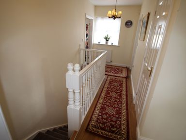 A hallway with a red patterned runner rug on wooden floor a white banister stairs a window with a plant and a chandelier at Vine Cottage in Charmouth