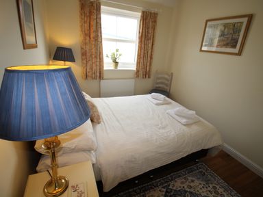 A bedroom with a double bed white linens two folded towels a window with floral curtains a chair a framed picture on the wall and two blue lamps at Vine Cottage in Charmouth