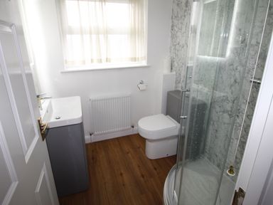 A bathroom with a shower, toilet, sink, radiator, and a window with curtains at Vine Cottage in Charmouth