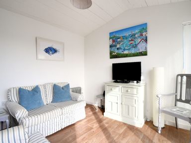 A living room with a striped sofa and blue cushions a cabinet with a television and two paintings on the walls at The Beach Hut in Lyme Regis
