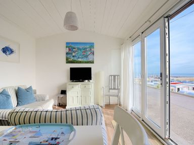 A sitting area with striped sofas a cabinet with a television and a chair next to large glass doors showing a view of a harbor at The Beach Hut in Lyme Regis
