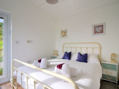 A bedroom with a metal bed frame white bedding purple pillows towels on the bed bedside tables with lamps and framed pictures on the walls at The Beach Hut in Lyme Regis