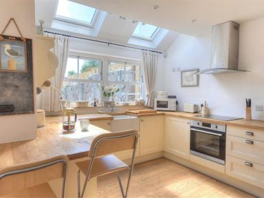 A kitchen with wooden countertops, cream cabinets, a sink under a window, a microwave, toaster, oven, and two chairs at a counter at Bethel Cottage in Lyme Regis