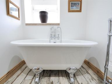A bathroom with a white clawfoot bathtub a window with a brown pot on the sill and wooden framed pictures on white walls at Bethel Cottage in Lyme Regis
