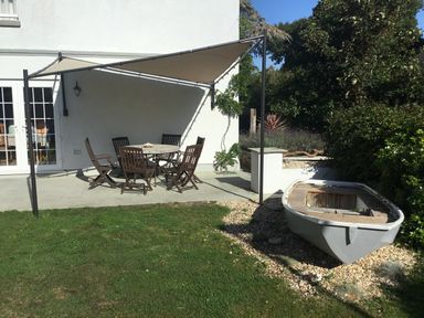 An outdoor patio area with a round wooden table and chairs under a shade sail and a small boat placed on gravel near the garden at Bethel Cottage in Lyme Regis