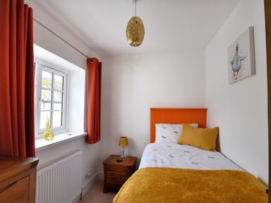 A small bedroom with a single bed orange headboard orange curtains and a wicker nightstand with a lamp at Sunnyside in Colyton