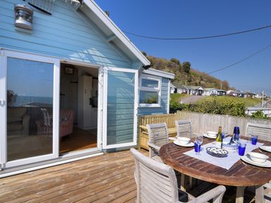 Outdoor wooden deck with dining table set with plates and glasses next to a blue house with open doors at Ammonite in Lyme Regis