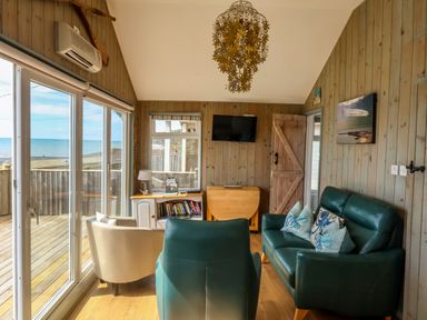 A sitting room with green sofas and a white armchair facing a television and sliding glass doors showing a sea view at Ammonite in Lyme Regis