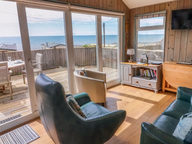 A living room with armchairs a cabinet a window and sliding glass doors showing a deck and ocean view at Ammonite in Lyme Regis