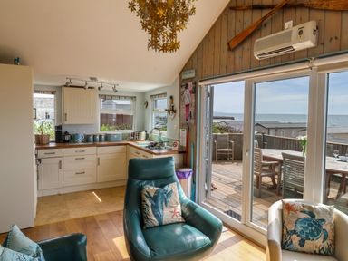 A kitchen with white cabinets and a seating area with green and white armchairs by sliding glass doors leading to a deck with outdoor furniture and a sea view at Ammonite in Lyme Regis