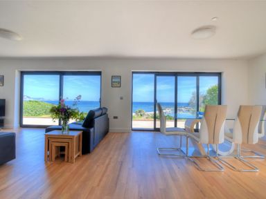 A living room with black sofas and a dining table with white chairs by large windows overlooking the sea at Harbour Heights Cottage in Lyme Regis