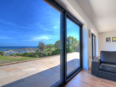 A living room with a black sofa and large glass sliding doors overlooking a patio and marina at Harbour Heights Cottage Lyme Regis